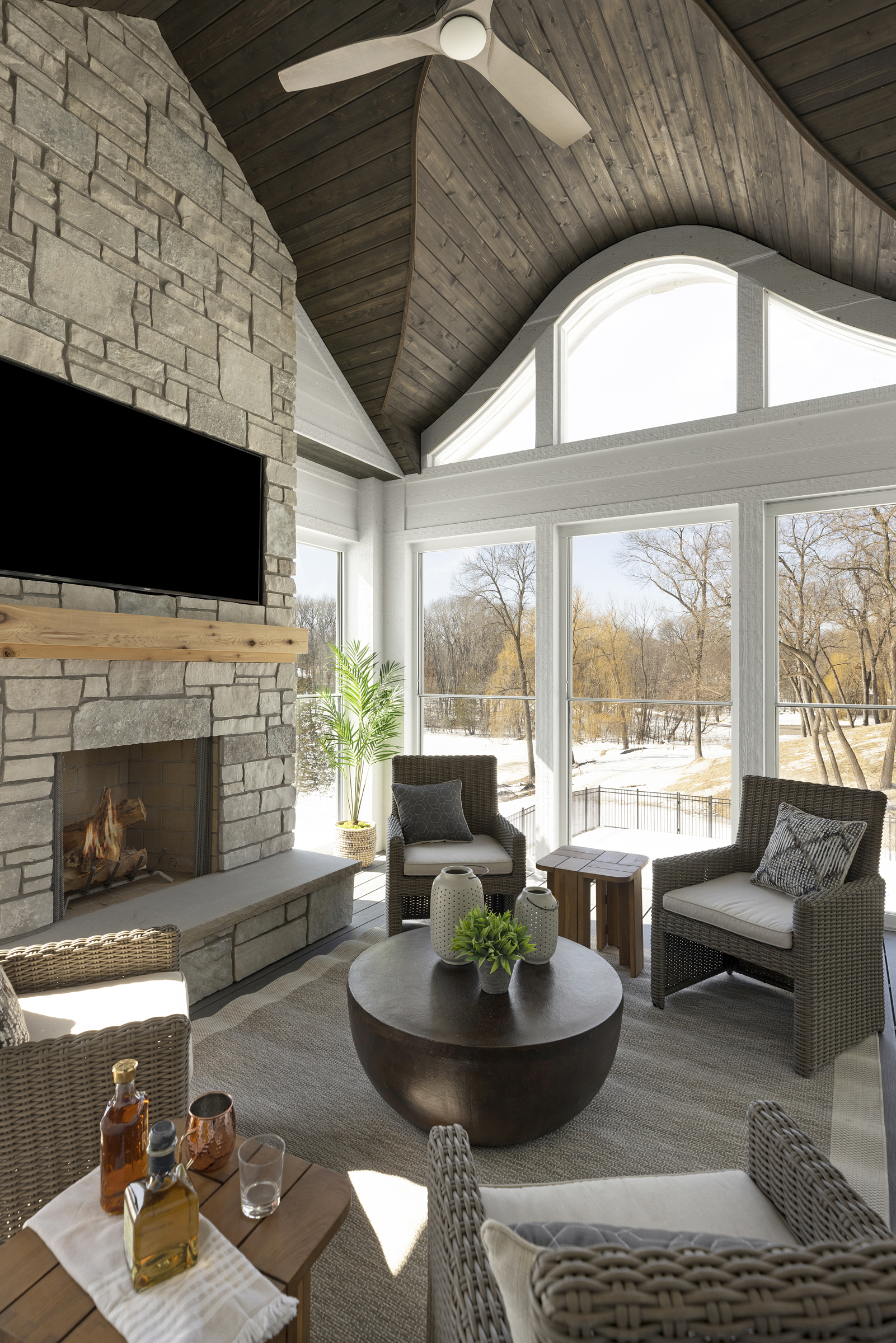 four season porch with arched ceiling detail, stone fireplace surround, floor to ceiling windows, four patio chairs, and a coffee table