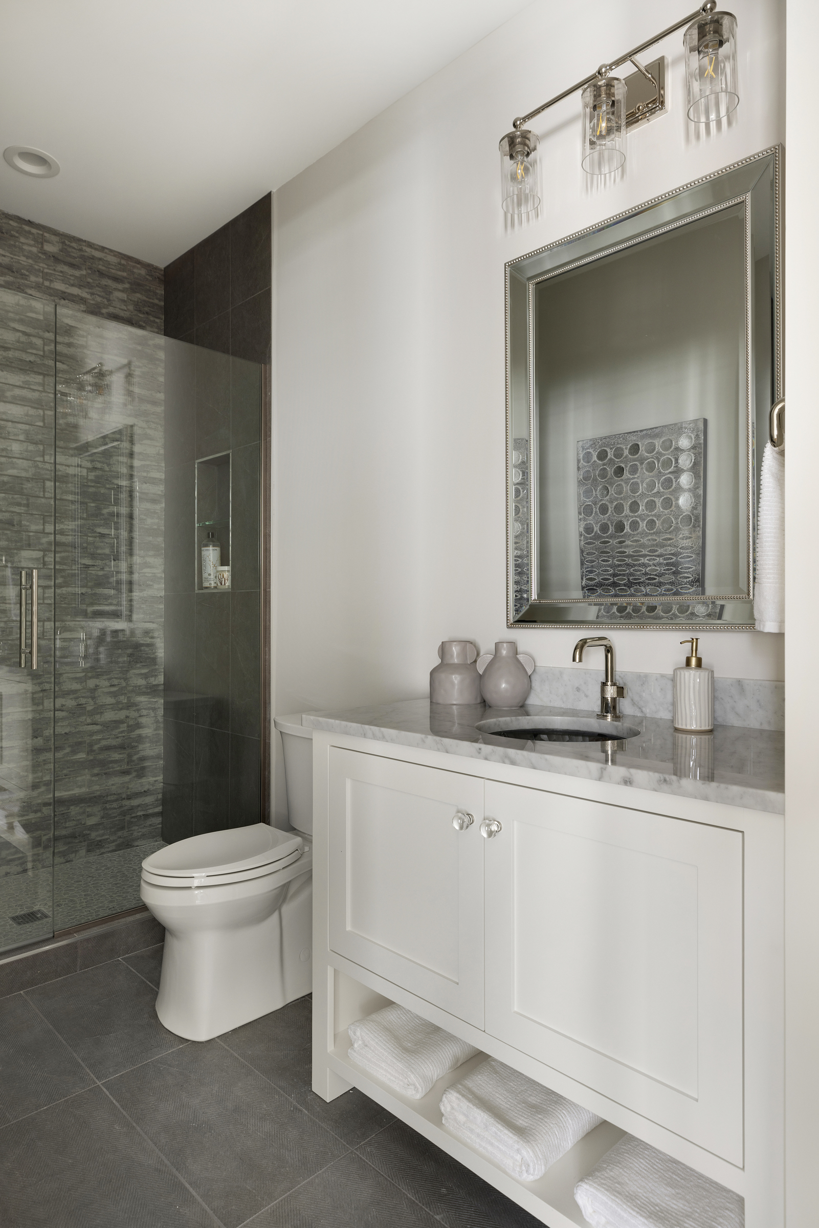 a bathroom with white walls, a glass shower on the left wall, a toilet in the middle, a white vanity on the right side with a sink and a mirror, and grey stone floors