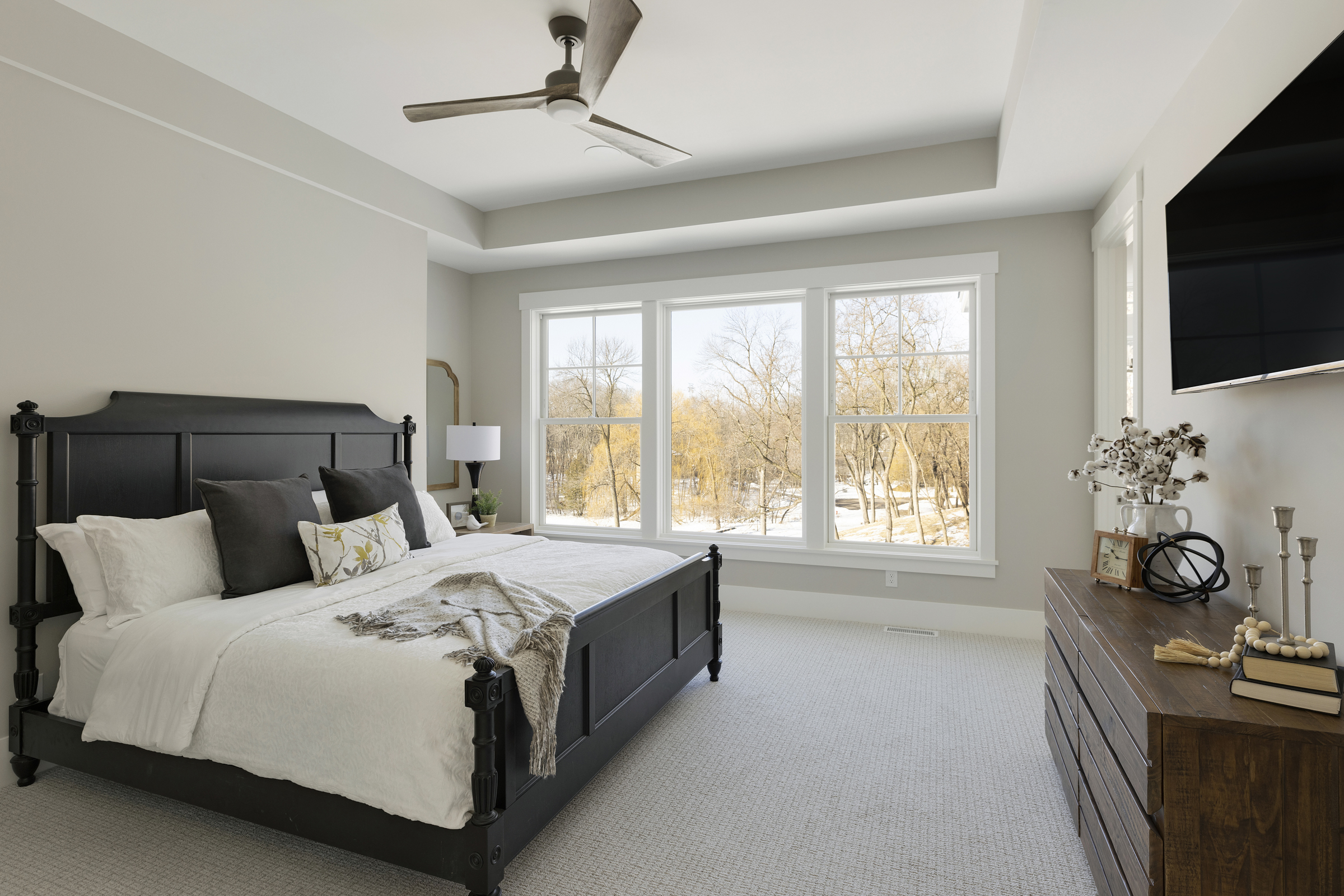 master bedroom with a king sized bed on the left side, 3 large windows on the back wall, and a brown dresser with a television hanging above it on the right