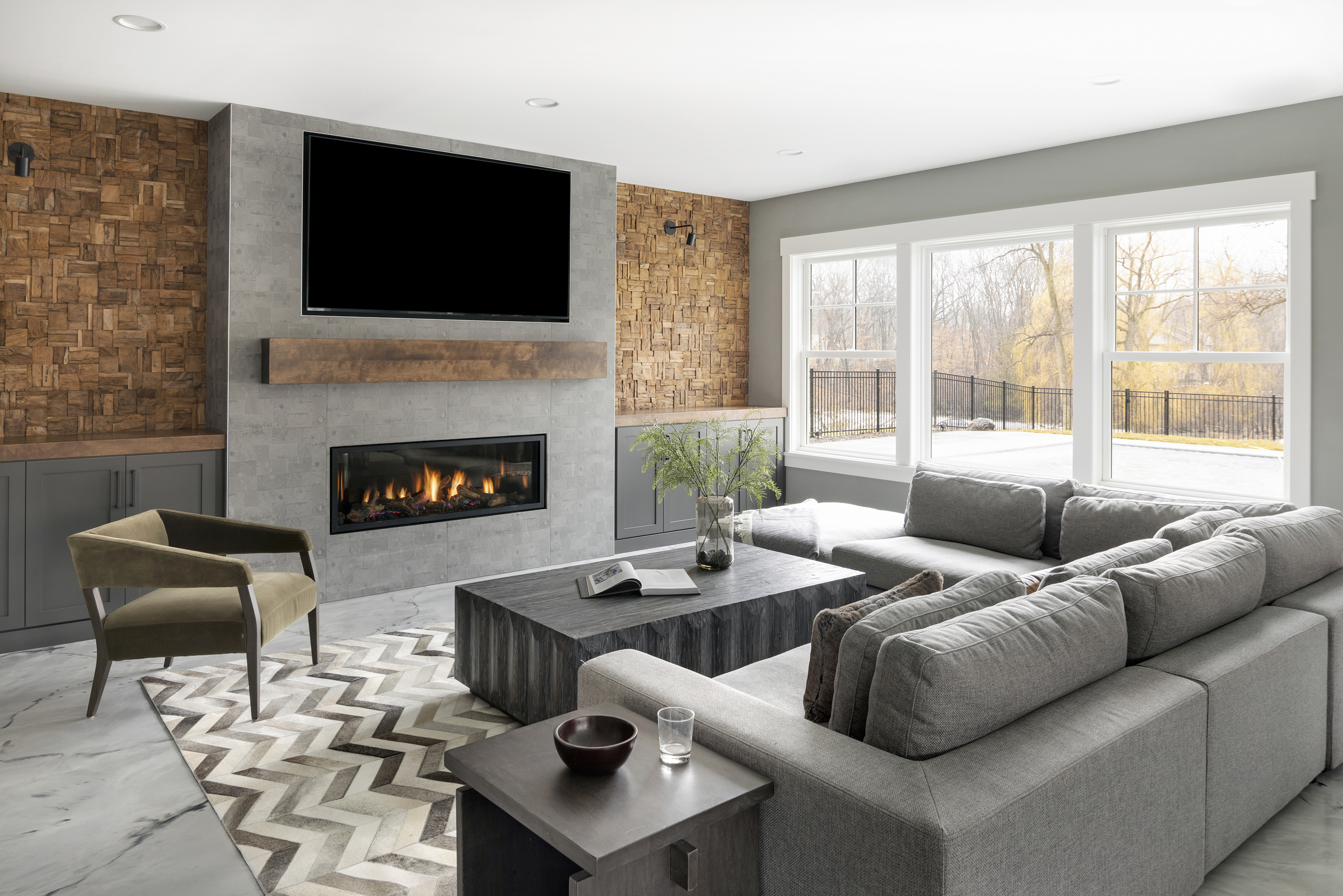 lower level living room with custom hand carved wood accent wall next to a custom stone fireplace surround with a reclaimed wood mantle, a grey sectional couch, a coffee table, and a chevron rug in the middle of the room