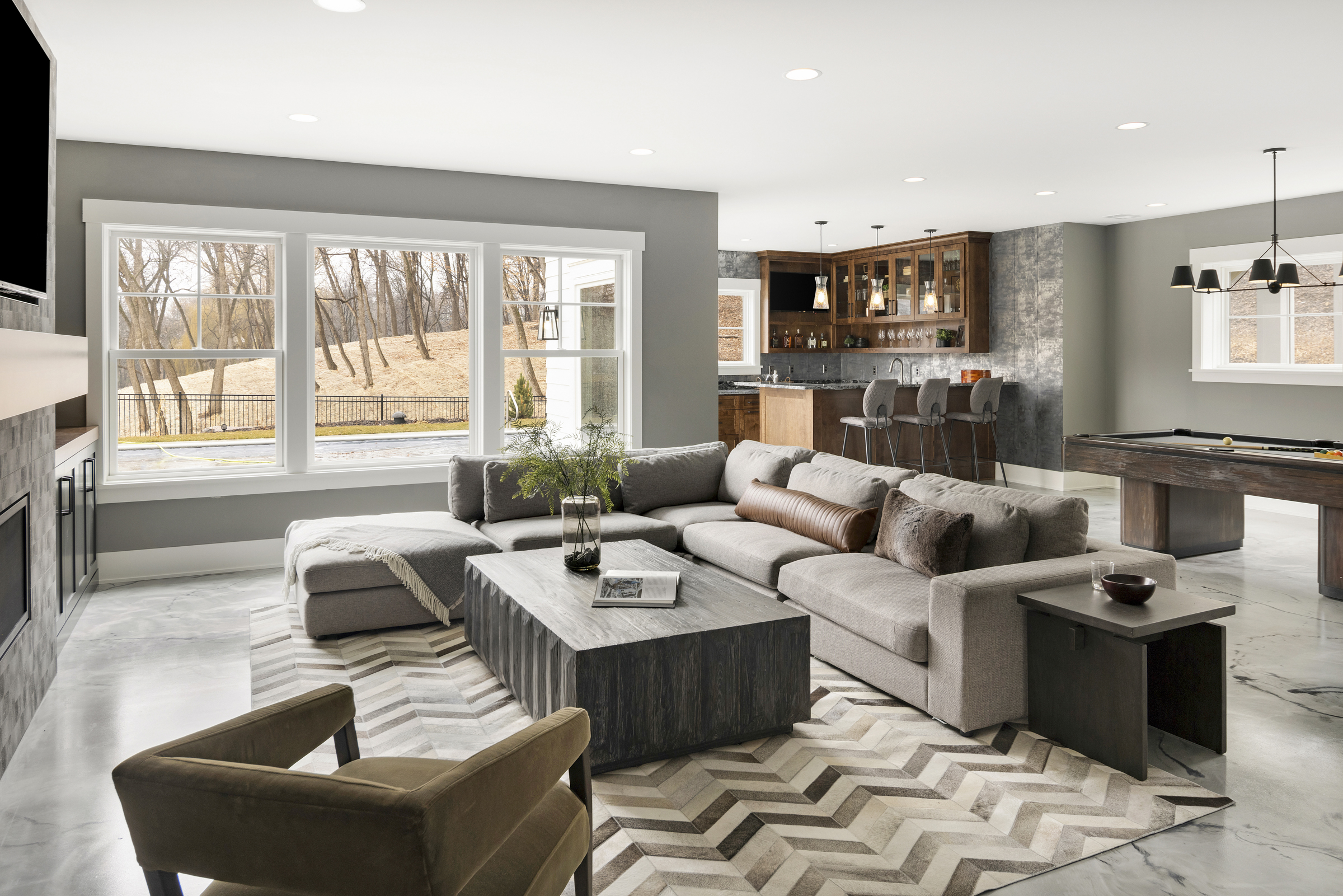 a lower level living room with a grey sectional couch, a dark coffee table, a chevron rug, and a view into the backyard through three large windows