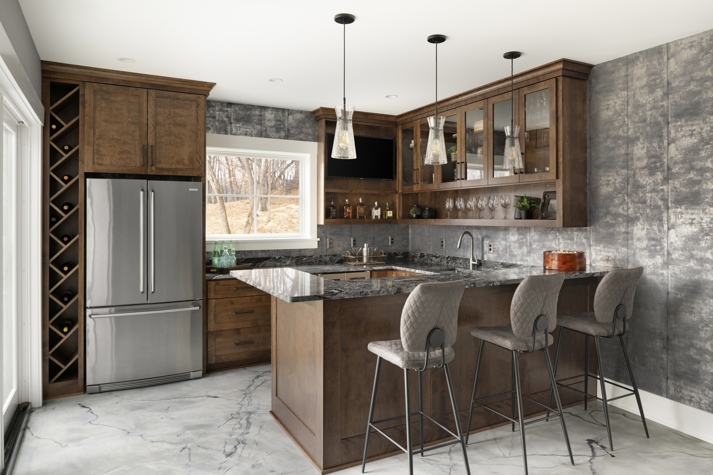 lower level bar in the corner of the room with wood cabinetry, a full sized fridge, a sink, three barstools at the counter, and metallic silver wallpaper on the walls
