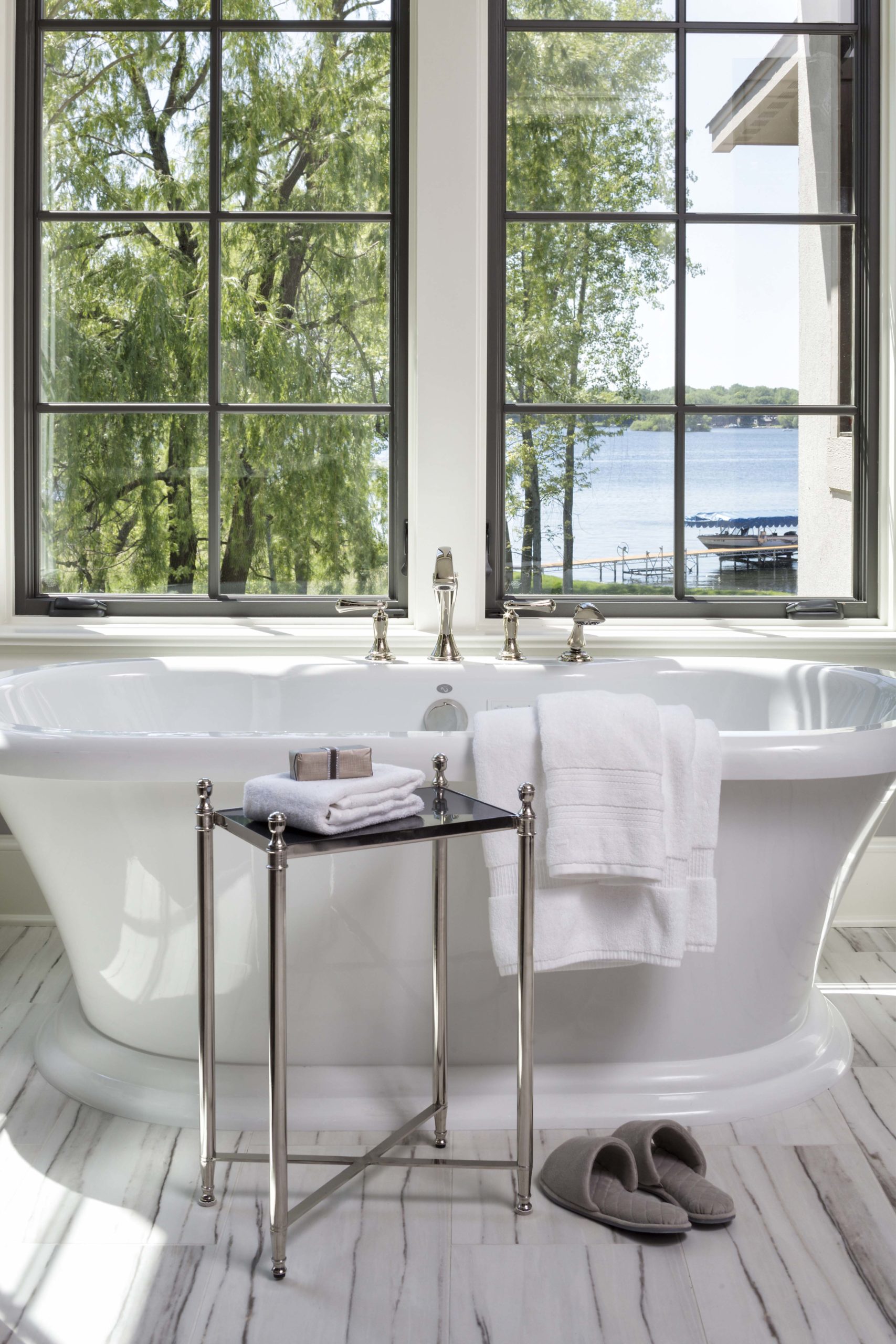white soaker tub on white marble floors with windows behind it looking out to the lake