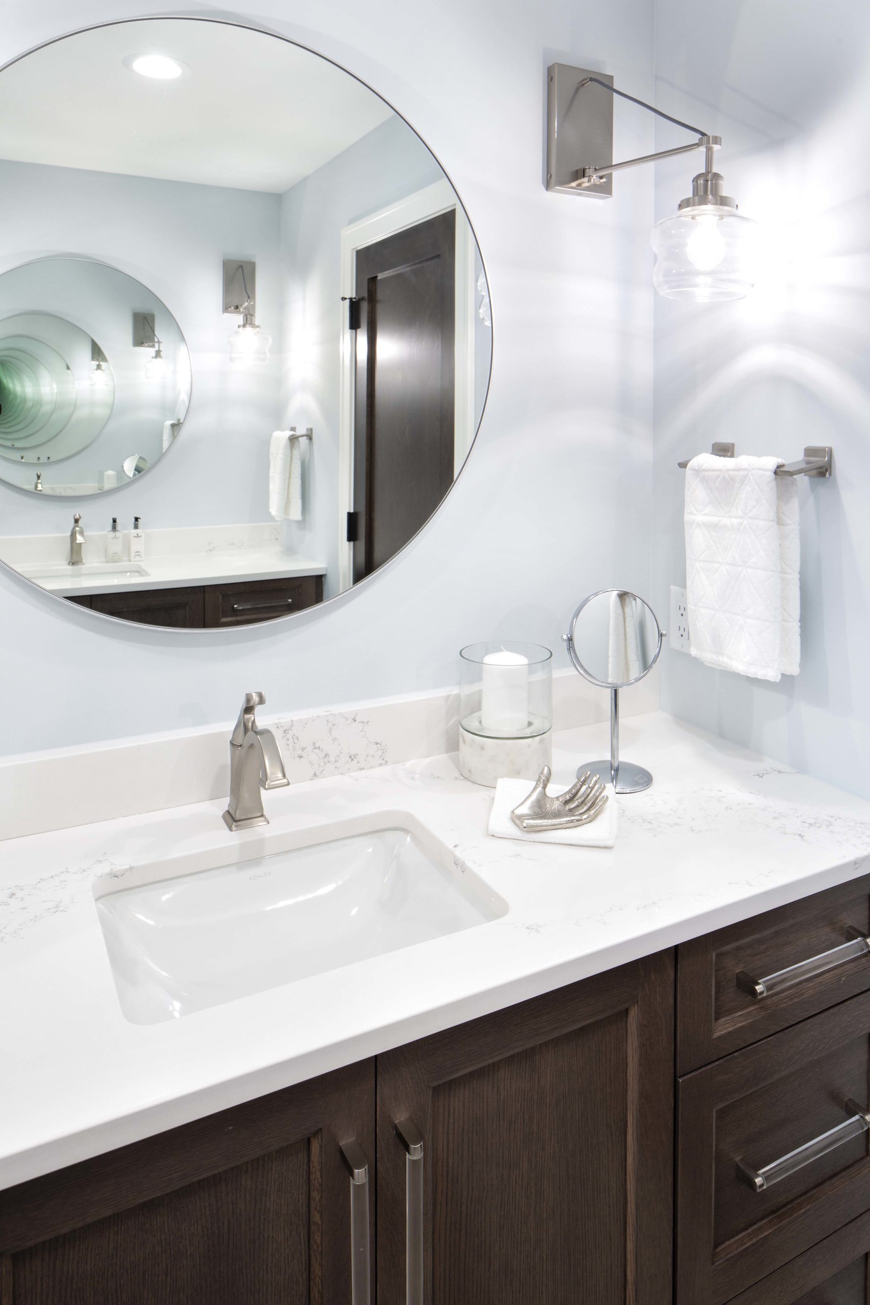 bathroom vanity with brown cabinets, white coutnertops, and a round mirror above
