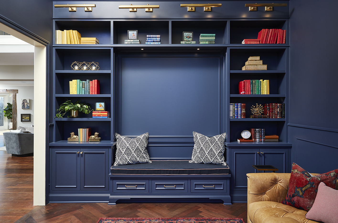 Library room with dark blue shelving filled with a rainbow of books, a brown leather couch sitting next to the shelves, and a custom piece of art on the wall