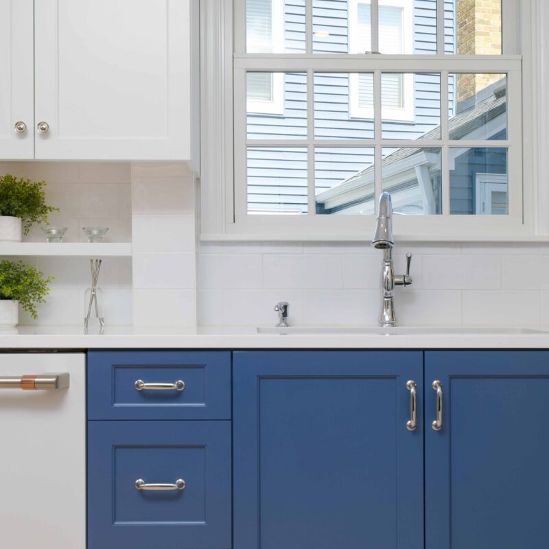 The Hampshire Drive kitchen remodel reveals a space with blue cabinets, a white countertop, and a modern faucet. A window above the sink offers a view of a neighboring house.