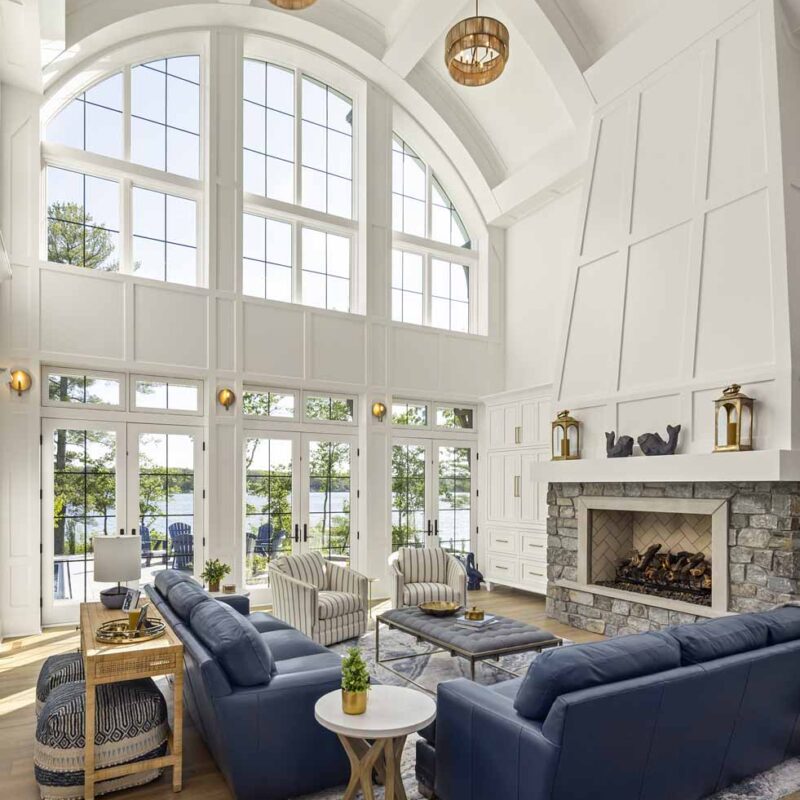 Spacious living room in this Nisswa Circle Lake home features high arched windows, blue sofas, a stone fireplace, and a wooden coffee table. Enjoy breathtaking views of trees and tranquil water just outside.