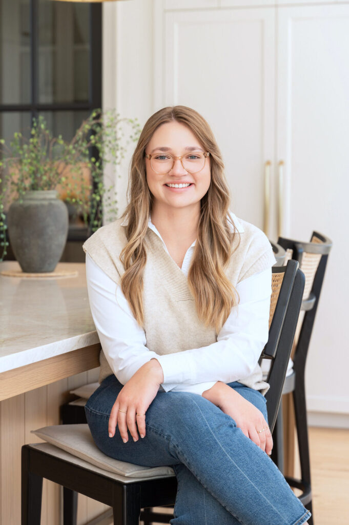 A person with long hair and glasses sits on a chair in a bright room thoughtfully curated by the Twin Cities interior design team, with a lush plant adding life to the table.